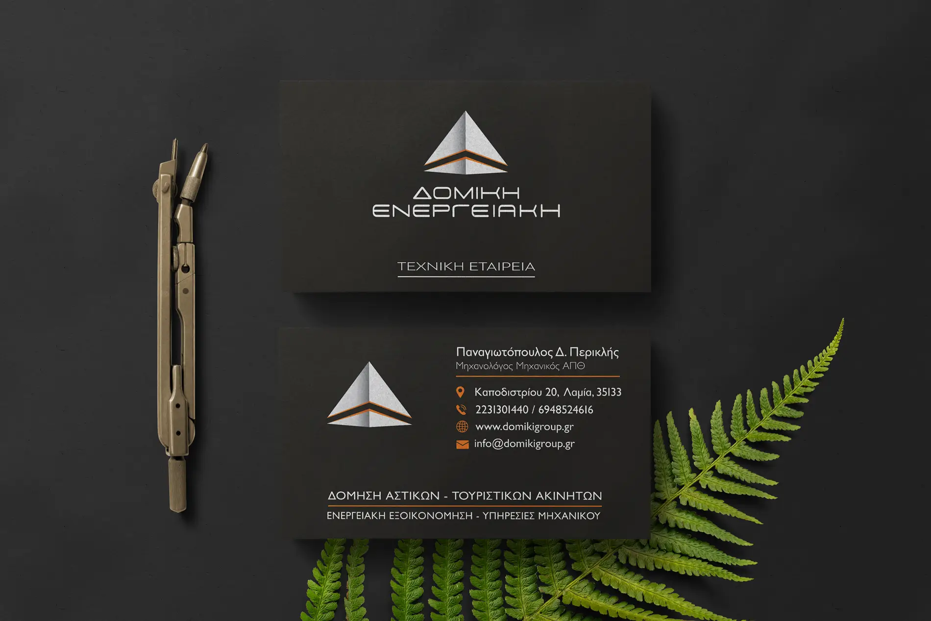 The dark business card shot from above on black paper — the logo face and the contact face, beside a pair of dividers and a fern